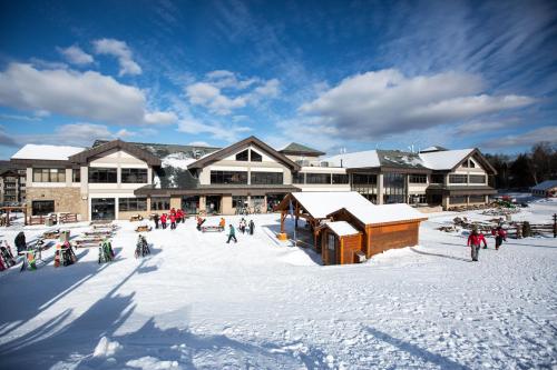 A snowy resort area with people walking, ski equipment, and a large building under a blue sky with clouds.