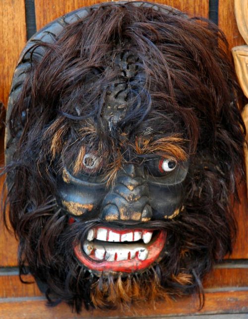 A detailed mask with a fierce expression, featuring dark hair, red eyes, and a prominent beard, mounted on a wooden background.
