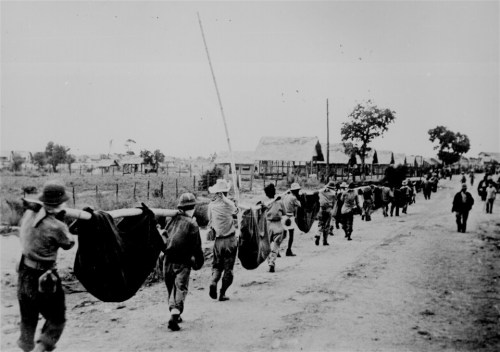 Black and white photo of soldiers carrying large objects along a dirt road, with houses and trees in the background.
