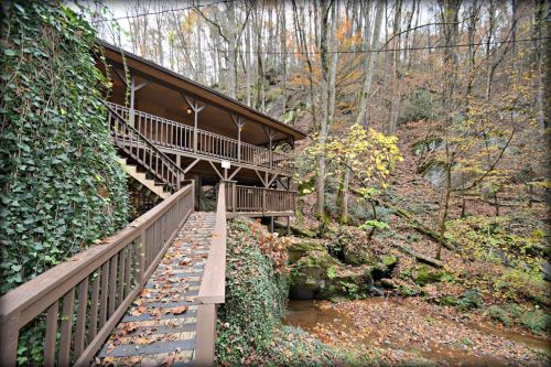 A wooden cabin with a staircase, surrounded by trees and autumn leaves, next to a small stream.