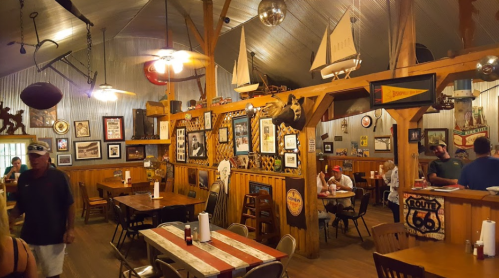 Cozy restaurant interior with wooden decor, vintage signs, and patrons dining at tables. Nautical theme with boats and memorabilia.