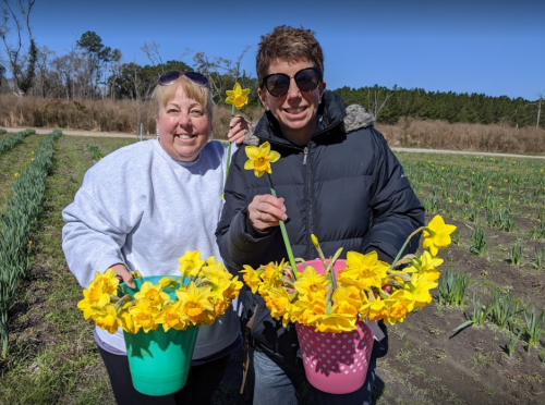 Two women smile while holding colorful buckets filled with bright yellow daffodils in a flower field.
