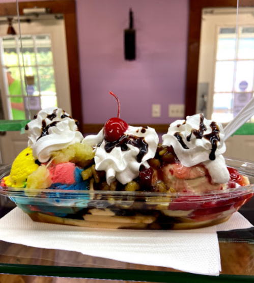 A colorful ice cream sundae with three scoops, whipped cream, chocolate drizzle, and a cherry on top, served in a dish.