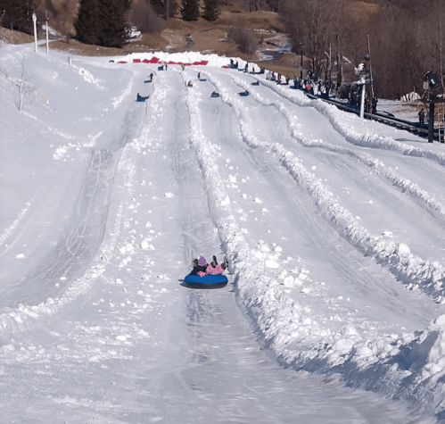 A snowy tubing hill with several people sliding down on inner tubes, surrounded by trees and a clear blue sky.