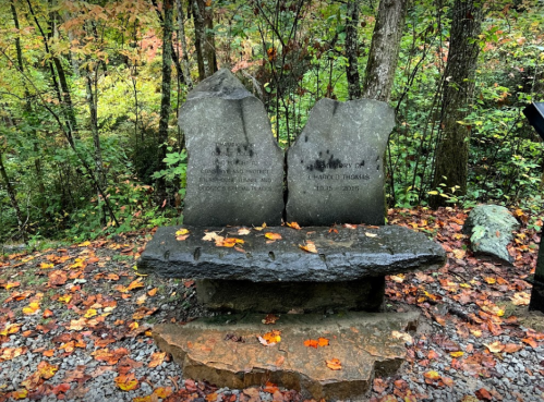 A stone bench in a forest, surrounded by autumn leaves, with inscriptions honoring a person named Harold Thomas.
