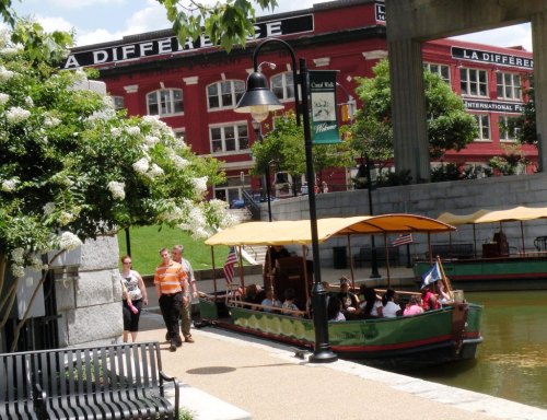 A scenic view of a canal with a boat and people walking along the path, surrounded by greenery and buildings.