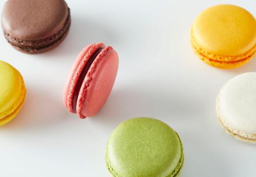 A colorful assortment of macarons in various flavors, including red, yellow, green, brown, and white, on a white background.