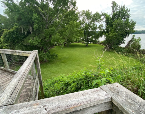 A wooden deck overlooks a lush green lawn bordered by trees and a body of water in the background.