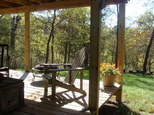 A wooden deck with an Adirondack chair and a potted plant, surrounded by trees and sunlight.