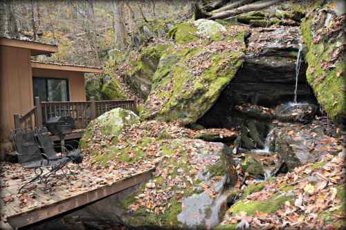 A cozy cabin with a deck beside moss-covered rocks and a small waterfall, surrounded by autumn leaves.