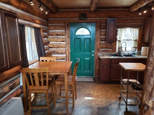 Cozy log cabin kitchen with a green door, wooden table, chairs, and a sink under warm lighting.