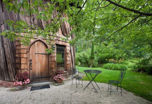 A rustic wooden building with an arched door, surrounded by greenery and a small outdoor seating area.
