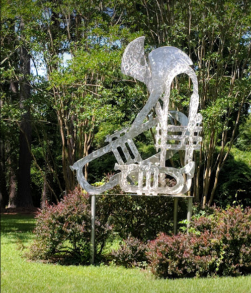 A metallic sculpture of a figure playing a saxophone, surrounded by greenery and bushes in a park setting.