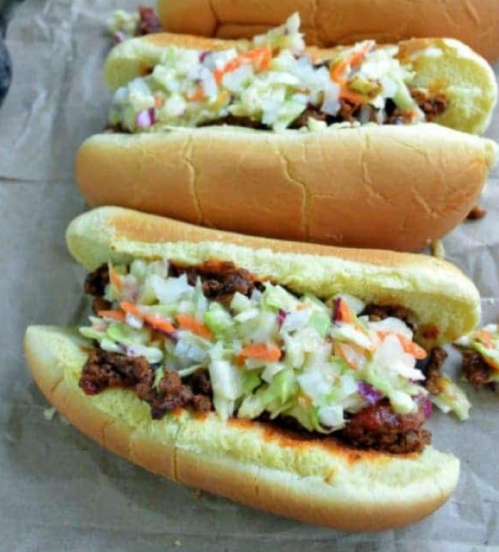 Three hot dogs filled with chopped meat and topped with coleslaw, served on a brown paper surface.