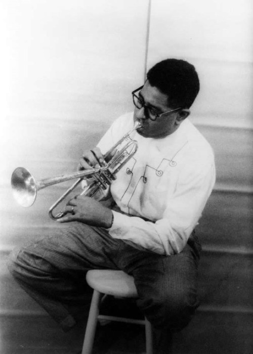 A musician playing the trumpet while seated on a stool, wearing glasses and a light-colored shirt.
