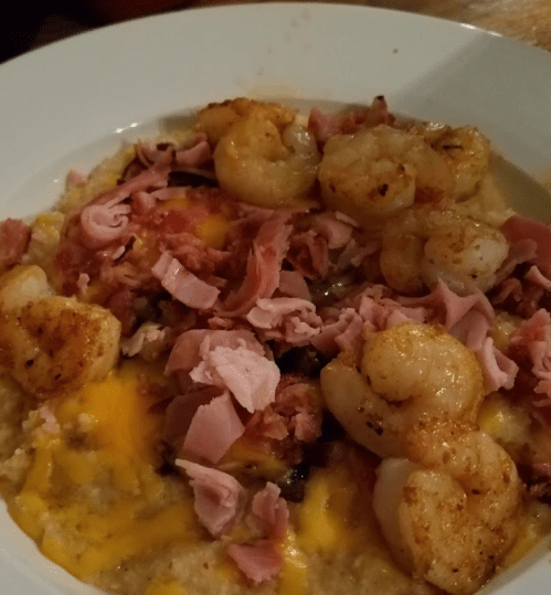A plate of creamy grits topped with shrimp, chopped ham, and melted cheese.