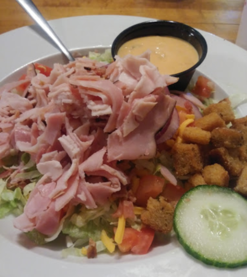 A salad topped with shredded ham, lettuce, tomatoes, cheese, croutons, and a side of dressing in a small cup.