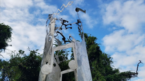 A tall, abstract metal sculpture with cut-out letters against a blue sky and green foliage.