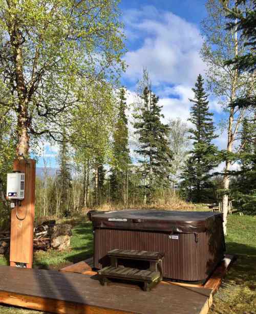 A hot tub on a wooden deck surrounded by trees and blue skies, with a small step for access.