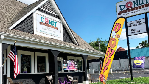 Exterior of Donut Distillery with a colorful sign, flags, and an American flag, promoting happy hour and trivia night.