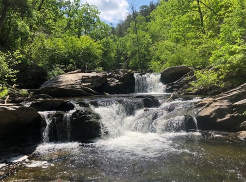 A serene waterfall cascades over rocks, surrounded by lush green trees under a bright blue sky.