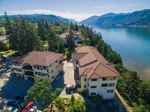Aerial view of a waterfront property surrounded by trees and mountains, with a clear blue sky and calm water.
