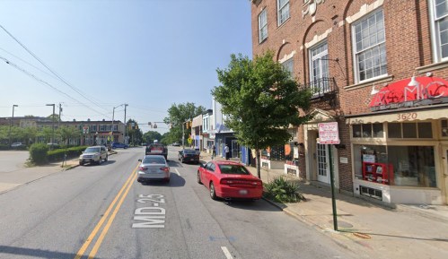 Street view of a small town with shops, parked cars, and a clear blue sky. Traffic signs visible on the road.