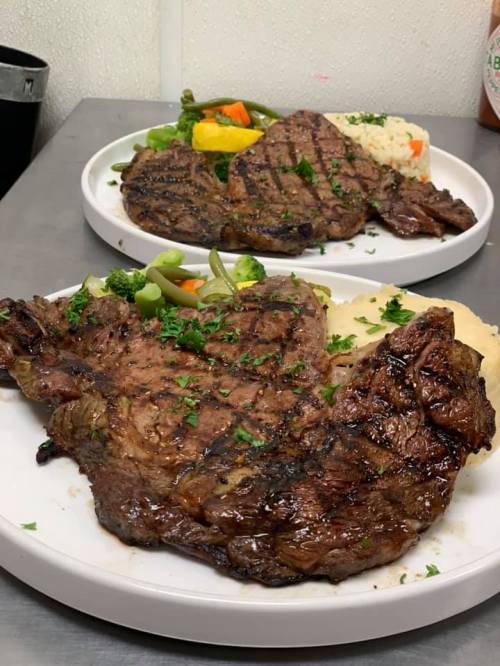 Two plates of grilled steak with garnishes, served with mixed vegetables and rice, on a stainless steel surface.