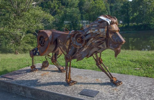 A metal sculpture of a lion, crafted from various scrap materials, positioned near a riverbank with greenery in the background.