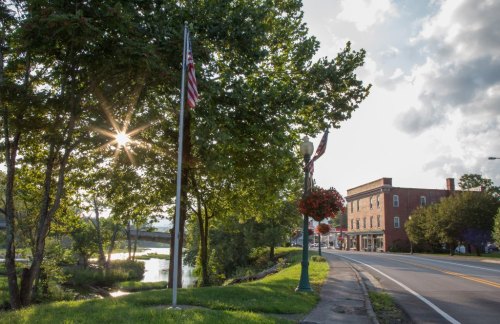 A scenic view of a tree-lined road with a river, flags, and a historic building under a sunny sky.