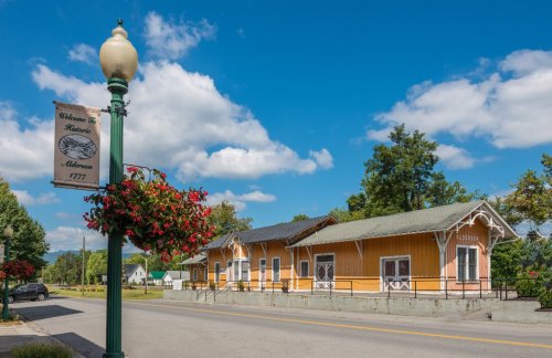 Historic buildings in a small town, with a street lamp and flower basket in the foreground under a blue sky.