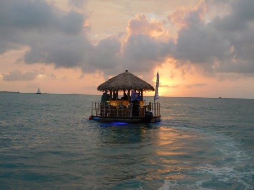 A floating tiki bar with a thatched roof on calm waters at sunset, with clouds and a distant sailboat.