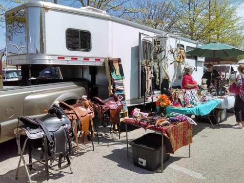 A vendor setup with saddles and accessories displayed outside a trailer, surrounded by shoppers at a market.