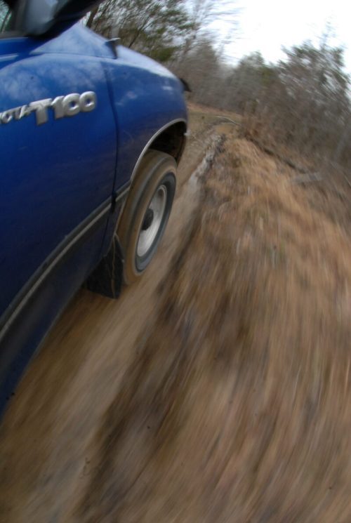 A blue truck driving on a muddy, uneven trail, captured in motion with a blurred background.
