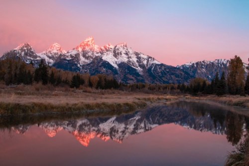 Sunset over snow-capped mountains reflecting in a calm river, surrounded by trees and golden grass.