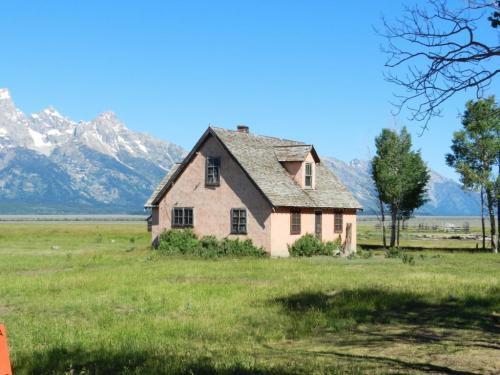 A weathered pink house surrounded by green grass, with mountains in the background under a clear blue sky.