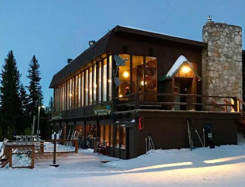 A cozy mountain lodge with large windows, surrounded by snow and trees, illuminated in the evening light.