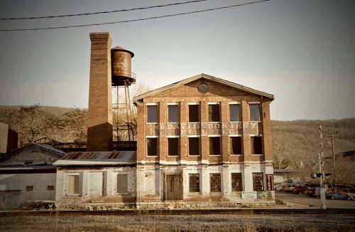 Abandoned brick factory building with a tall chimney and water tower, set against a hilly landscape.