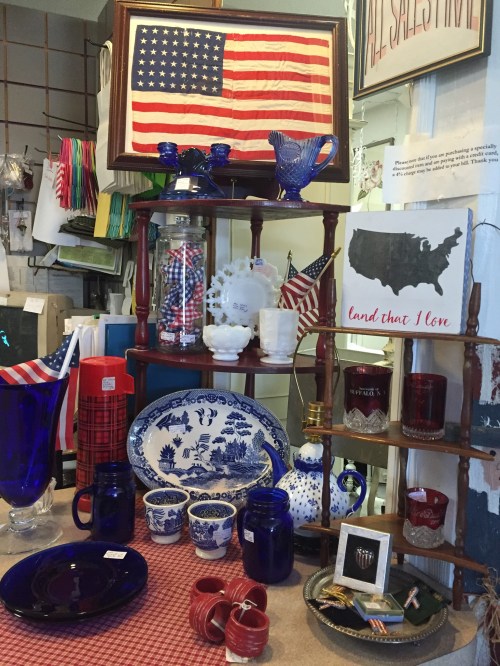 A display of patriotic decor and tableware, featuring a U.S. flag, blue glassware, and various red, white, and blue items.