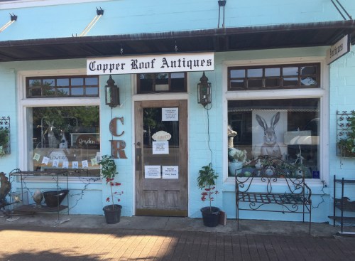 Exterior of Copper Roof Antiques shop, featuring a blue facade, large windows, and decorative plants.