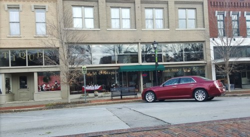 A red car parked on a street in front of a row of shops with large windows and a green awning.