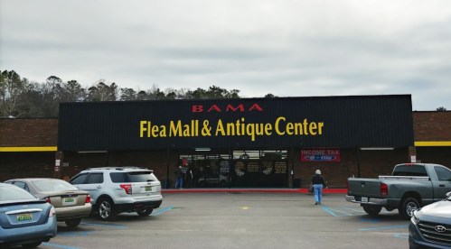 Exterior of BAMA Flea Mall & Antique Center, featuring a large sign and parked cars in front. Overcast sky in the background.