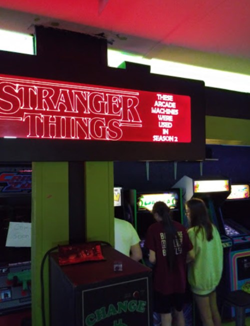 A neon sign reads "Stranger Things" above arcade machines, with two people playing games in a retro arcade setting.