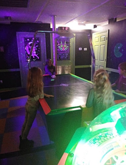 A group of children playing air hockey in a colorful arcade with neon lights and a scoreboard.