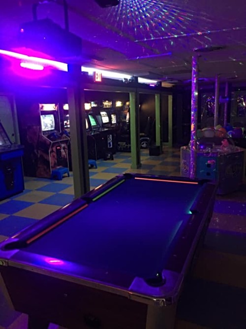 A dimly lit arcade room featuring a pool table and vintage arcade games under purple and blue lighting.