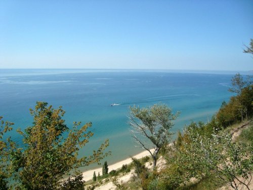 A serene view of a blue lake with a sandy shore, surrounded by trees under a clear blue sky.