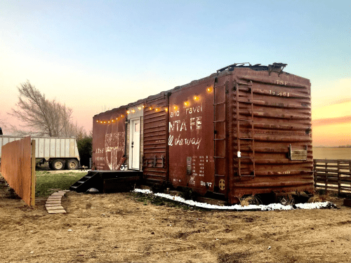 A rustic shipping container home with string lights, surrounded by a wooden fence and grassy area at sunset.