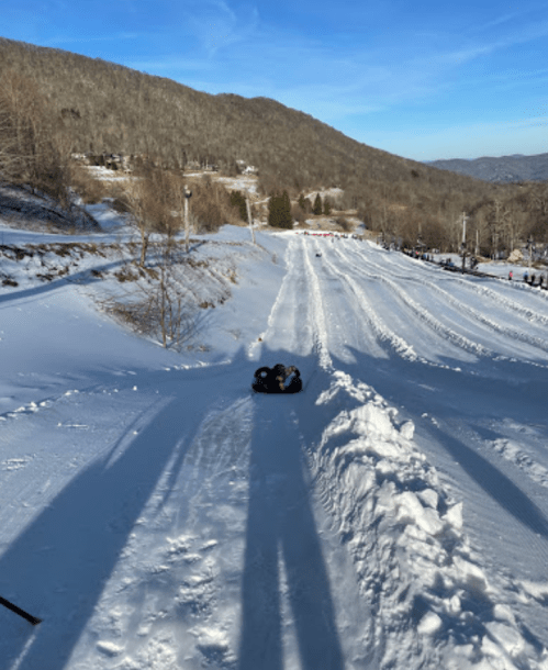 A snowy hill with tubing tracks, showing a clear blue sky and distant mountains in the background.