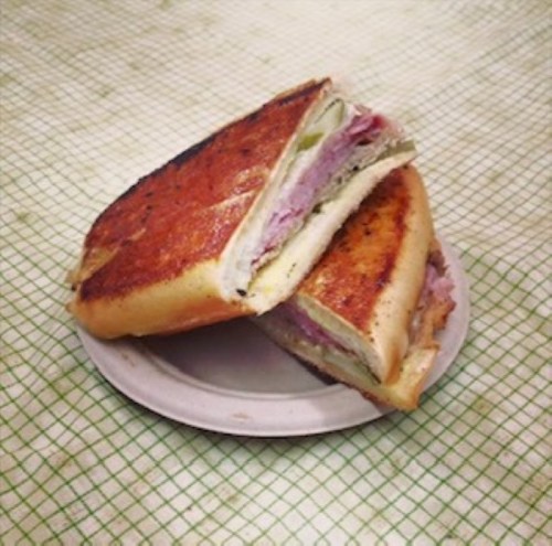 A toasted sandwich with layers of ham and pickles, served on a white plate against a checkered background.