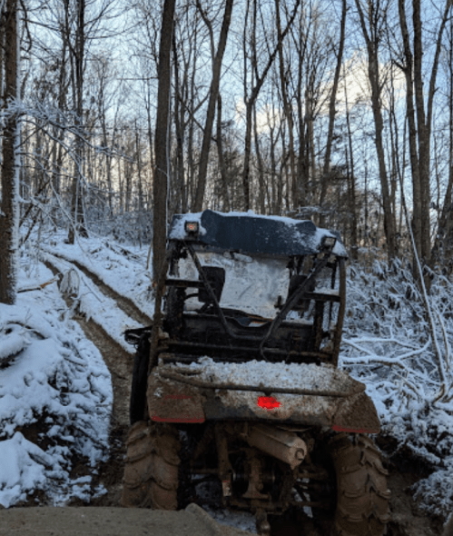 A muddy all-terrain vehicle navigates a snowy forest trail surrounded by trees.